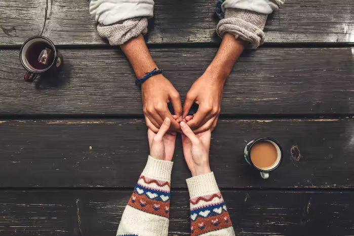 Two people holding hands across a wooden table with coffee cups on it, viewed from above.