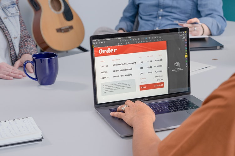Person using a laptop to update order form templates for their music store with an acoustic guitar in the background and two customers sitting at the table.