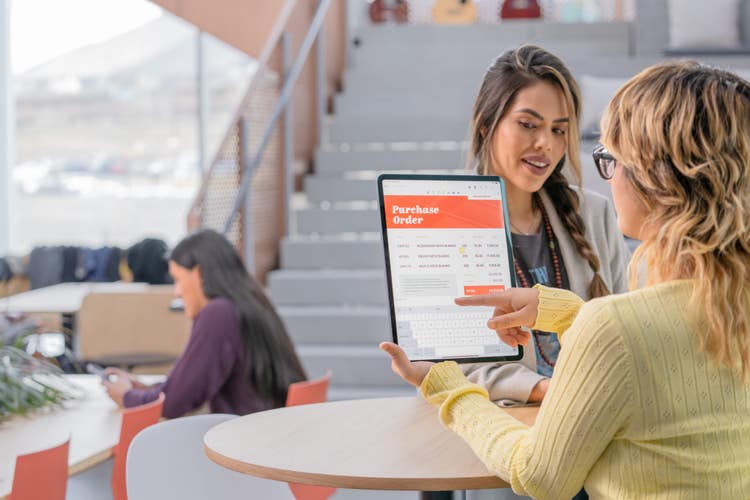A woman standing across from another woman at a high-top table uses invoicing software on her tablet.