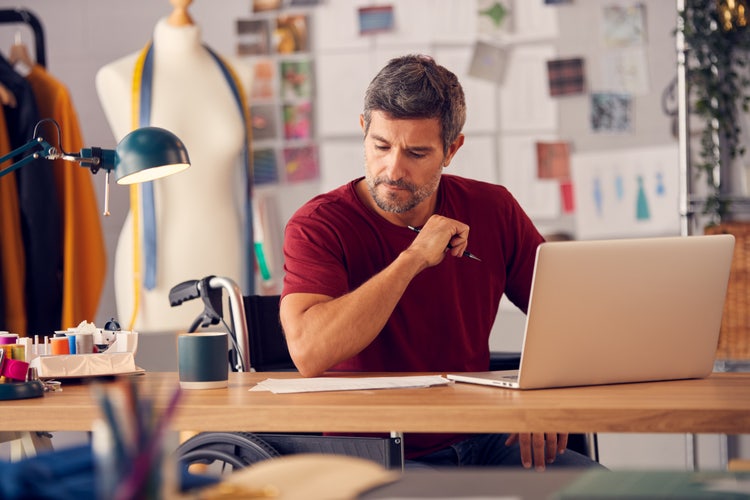 A man sits at a desk with his laptop and paper copies of common business forms