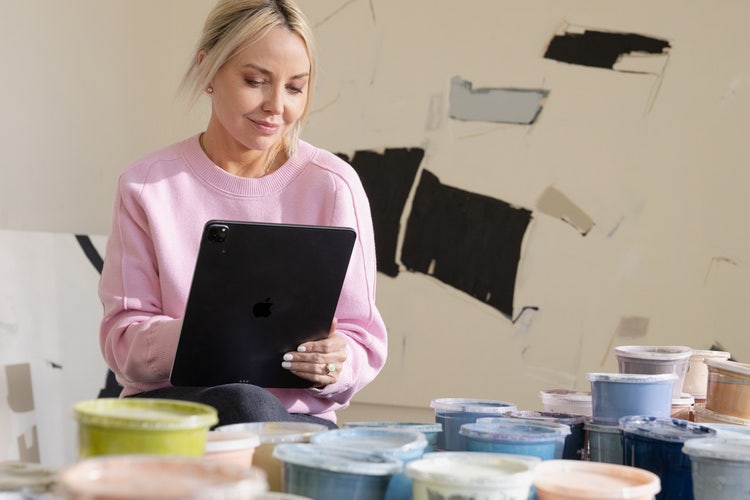 Woman in a pink sweater sitting in an art studio surrounded by paints using a tablet to write a business cover letter template.