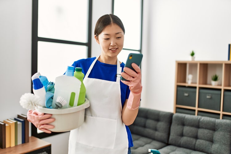 A woman holding a bucket of cleaning supplies with one arm holds a smartphone in her other hand and creates a cleaning invoice.