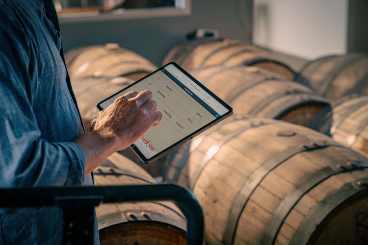 A person fills out an invoice on their tablet with wooden barrels in front of them.