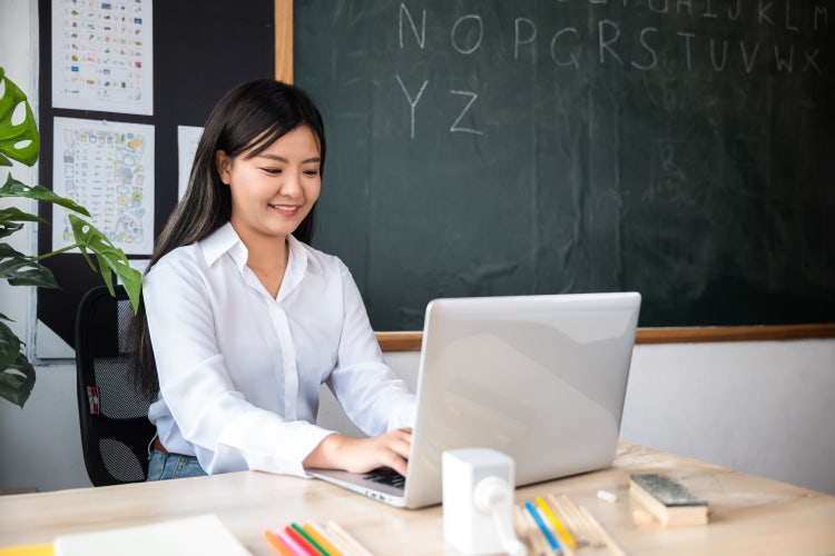 A woman sitting at a desk in a classroom creates an invoice for tutoring on her laptop.