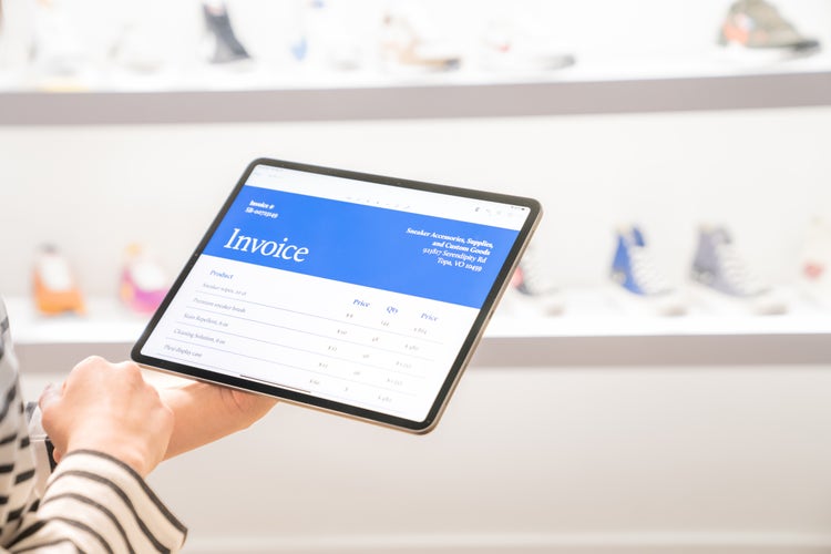 A woman holds a tablet with an invoice displayed on it, and in front of her are shelves of shoes.