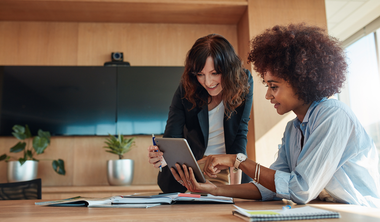 A woman standing holds a tablet up to another woman sitting at a desk.