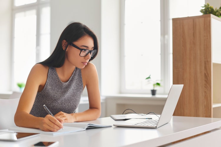 A woman sitting at a table writes in a notebook while looking at her laptop to calculate late fees on overdue invoices.