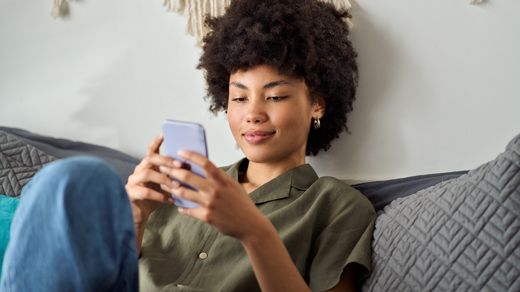 A woman sits on a couch, holding a smartphone.