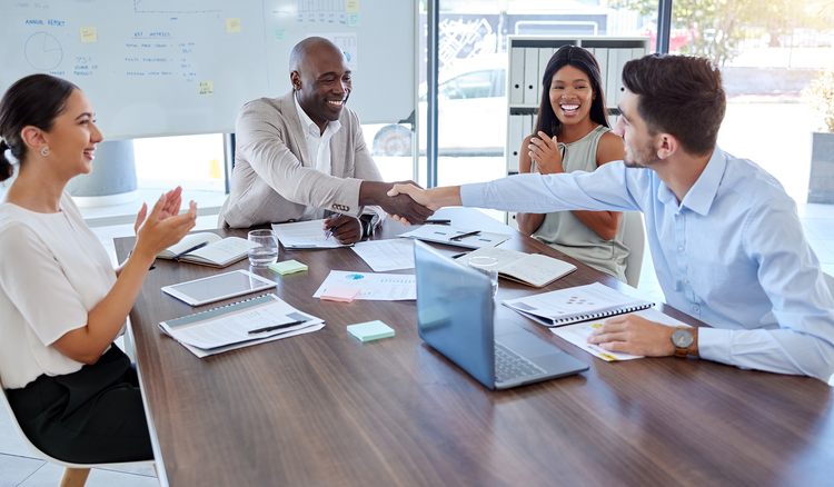 Four people, each with notes on the table in front of them, sit at a table in a conference room, where two people shake hands.