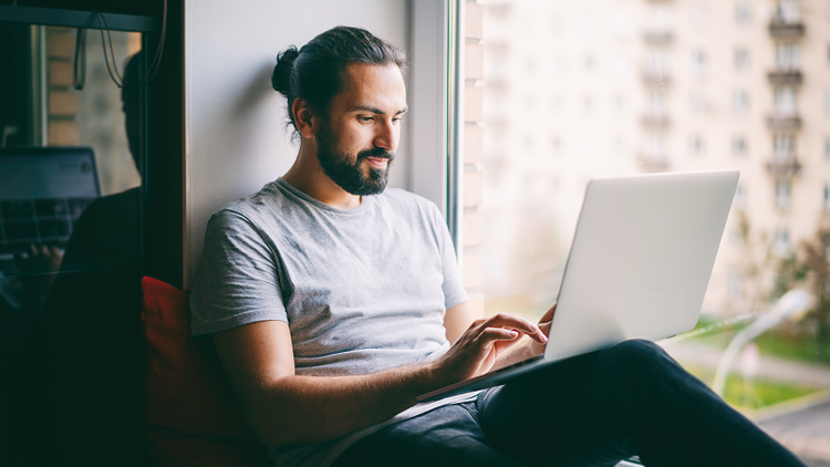 A man sitting on a windowsill types on his laptop.