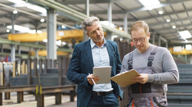 Two people standing in a factory and filling out some forms.