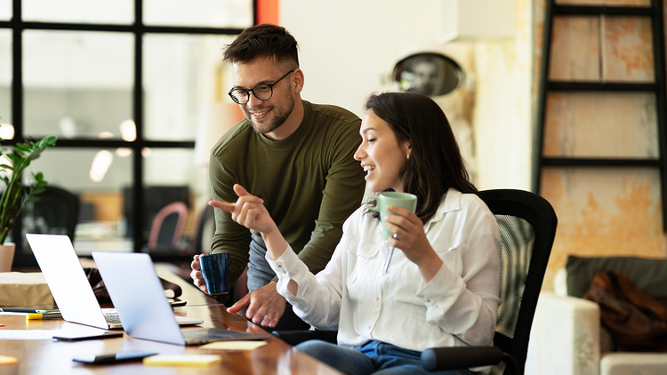 A man stands over a desk where a woman sits, pointing at a laptop.