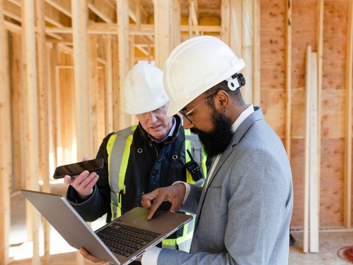 A contractor in a jumpsuit and a client in a suit looking over construction plans on a work site