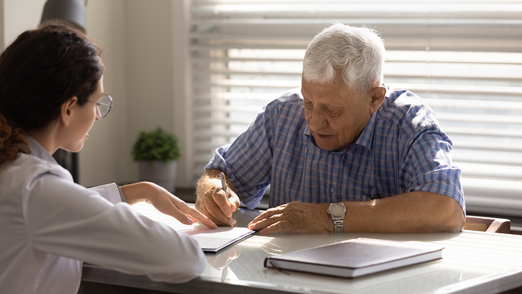 A man signs a form with a woman in a lab coat sitting opposite him.