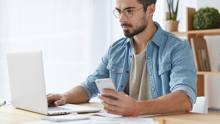 A man sitting at a desk holds a smartphone in his left hand and types on a laptop with his right.