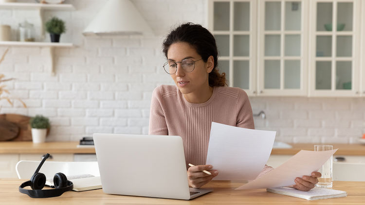 Woman holds two papers while looking at a laptop to review a mortgage note.