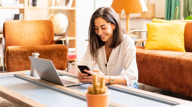 Woman with black hair in a white shirt using her laptop and phone while sitting at a coffee table