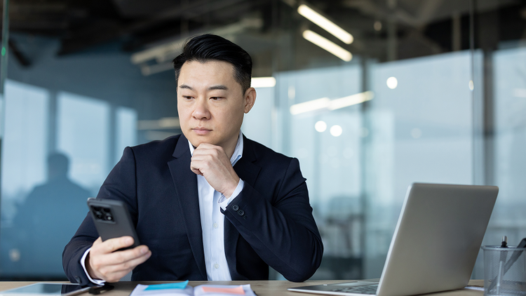 A man sitting at a desk with a laptop in front of him looks at his smartphone.