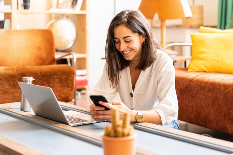 Woman with black hair in a white shirt using her laptop and phone while sitting at a coffee table