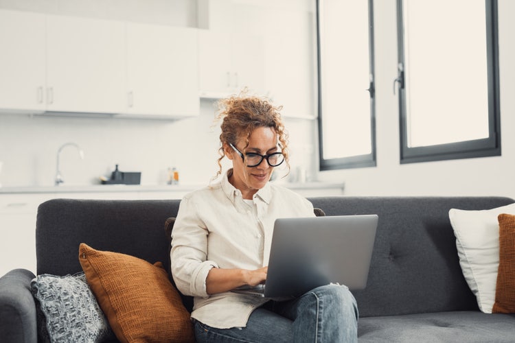 A woman wearing glasses sitting on a couch in a bright room and typing on a laptop.
