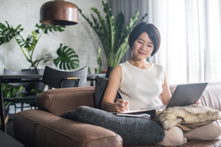 A young woman working on a couch in a home or office space with natural light and greenery. She has a laptop on her lap while writing in a notebook next to her.