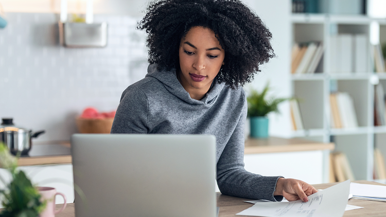 A woman sitting at a table holds a sheet of paper in one hand and looks at her laptop computer.