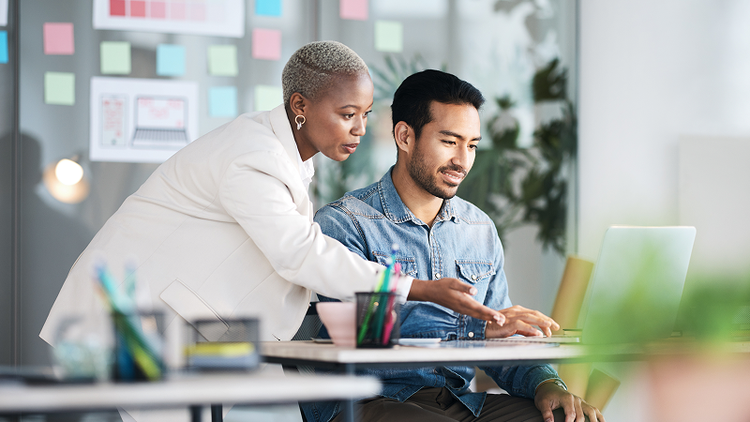 A woman standing over a man's shoulder gestures at a laptop on a table.