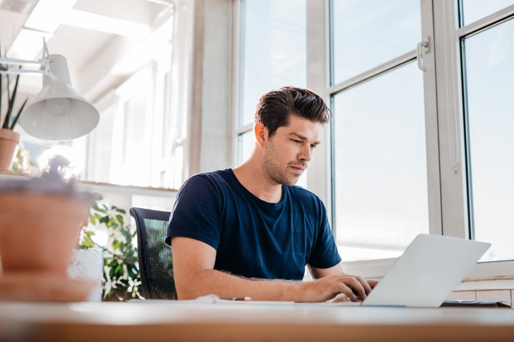 Focused young man working on a laptop in a bright modern office with large windows and indoor plants.