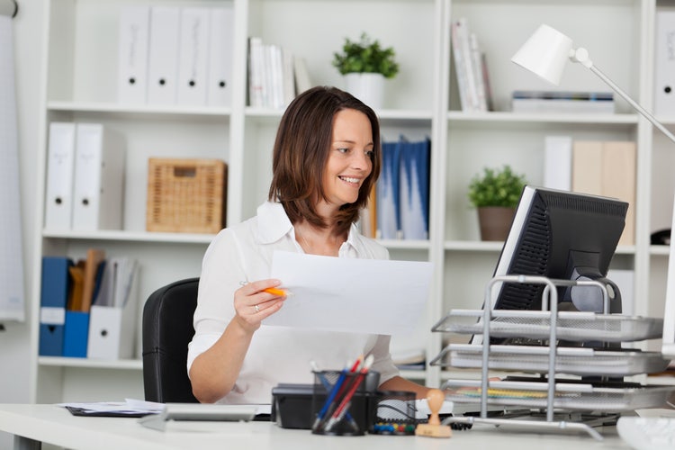 A woman in an office holds a printed version of a PowerPoint with notes and compares it against the presentation on her computer.