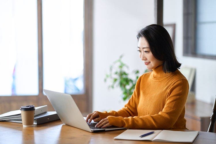 A woman wearing an orange sweater smiles while working on a laptop to save and print a PowerPoint as a PDF with notes.