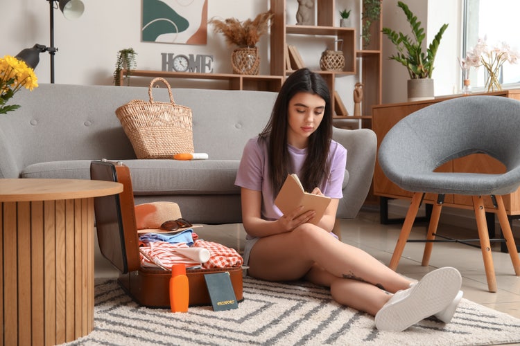 A woman sitting on a living room floor next to a suitcase writes down a cruise packing list in her notebook.