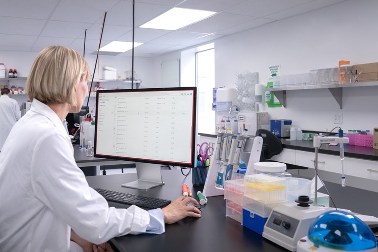 A woman in a white coat works at a computer in a laboratory, learning how to save a Google Doc as a PDF.