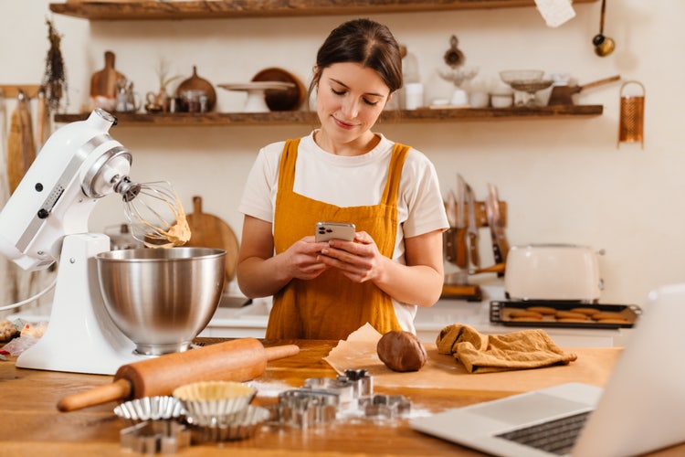 A woman is digitizing a recipe on a mobile phone app while baking in the kitchen.