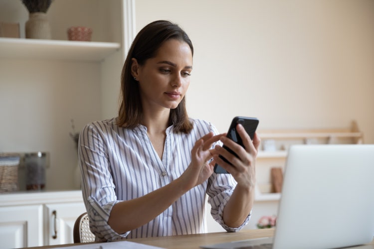 A woman sitting at a table with a laptop in front of her is holding a smartphone.