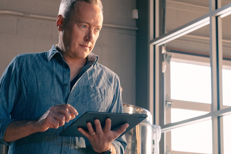 A person in a blue button-up shirt standing in front of a distillery tank and garage doors working on a fact sheet template PDF using a tablet.