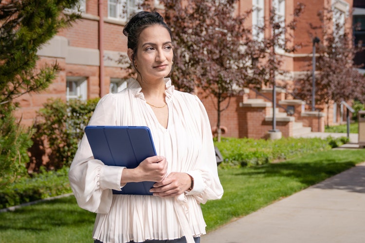 A woman holding a large tablet in a navy-blue case walks around a university campus with red brick buildings, looking for a spot to sit and read about how to ask a professor for a letter of recommendation.