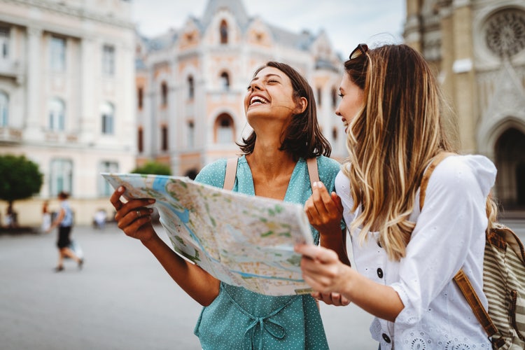 Two women standing in the middle of a town square laugh while holding a map and making a travel budget.