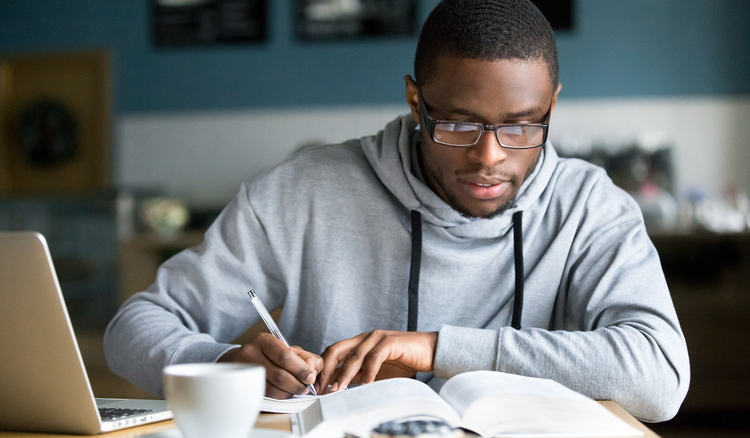 A man sitting at a desk with a laptop and a book in front of him writes in a notebook.