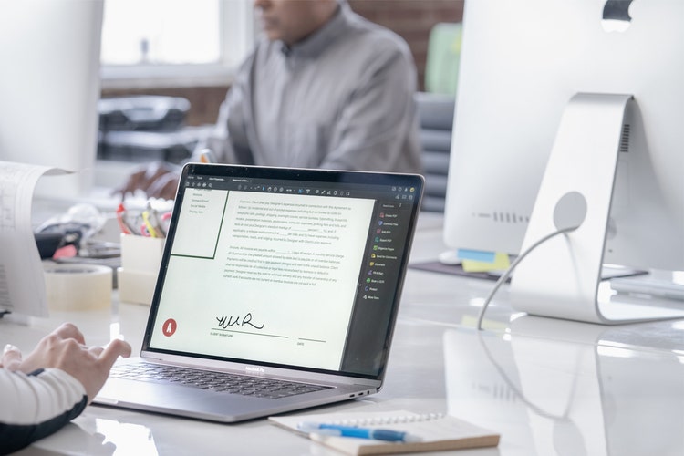 Person working in an open office space at a large, shared desk with laptops and monitors getting a signed document ready to scan.
