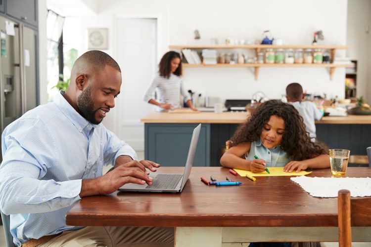 A father and daughter sitting at a kitchen table. The father is working on a laptop while his daughter sits next to him coloring a picture.