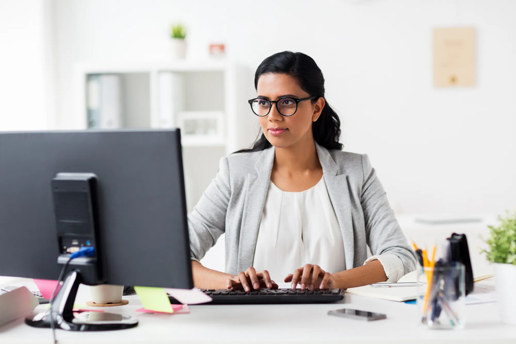 A woman sitting at a desk writes and types on a PDF using her PC.