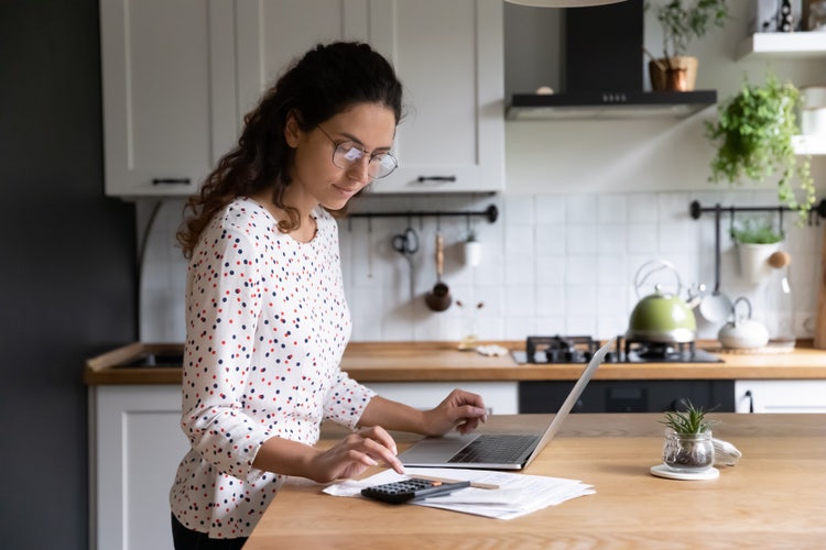 A woman standing over a kitchen island table creates a weekly budget template on her laptop.