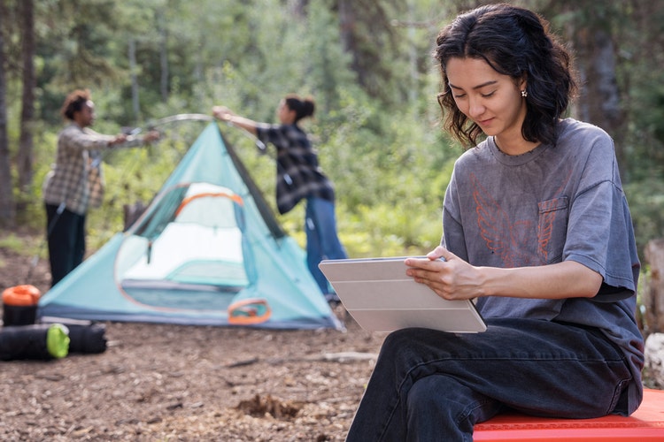 Casually dressed woman sitting at a campsite with friends while working on her tablet learning how to make a time blocking template.