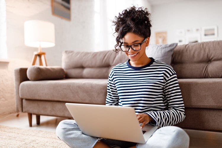 A young woman wearing a striped shirt sitting on the floor using a laptop to download a PDF of a newspaper.
