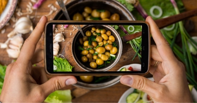 Two hands holding a mobile phone with the camera turned on and ready to take a photograph of a robust spread of food on the table below.