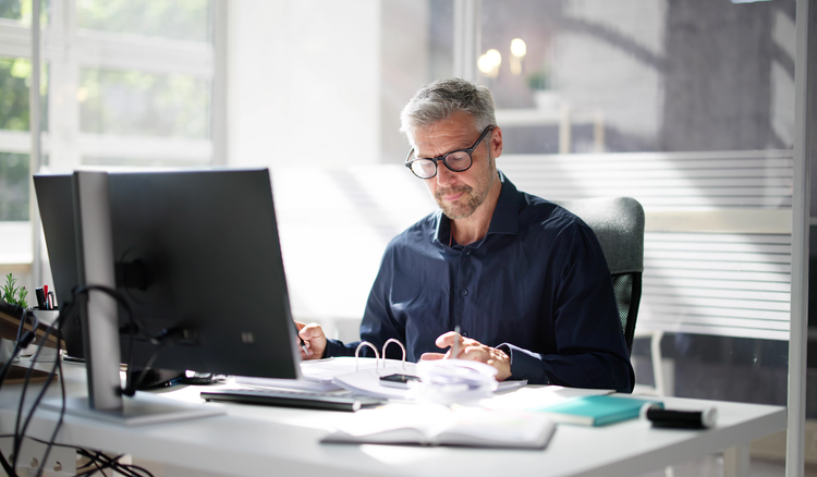 A man sitting at a desk looks at a binder full of documents.