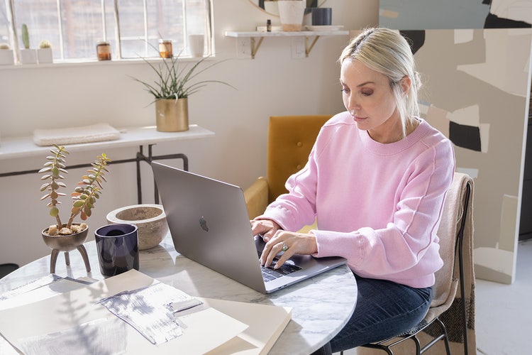 Woman in a pink shirt sitting at a marble-top table working on a laptop to create a job reference template.