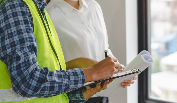 Two people stand next to each other with one holding a clipboard and writing on a piece of paper.