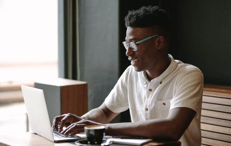 A man sitting in a coffee shop sits at a table and types on his laptop to create a cover letter for his resume.