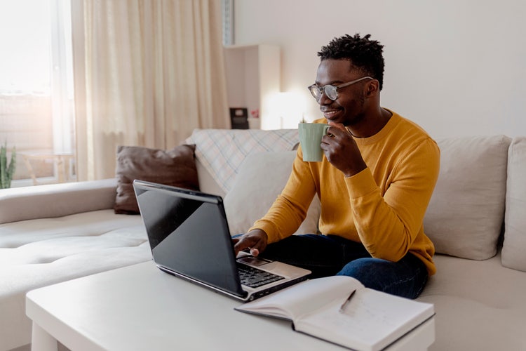 A man in glasses and a mustard sweater sits on a beige couch, drinking from a green mug and working on a laptop to write a cover letter for a teaching job with an open notebook on the white coffee table in front of him.